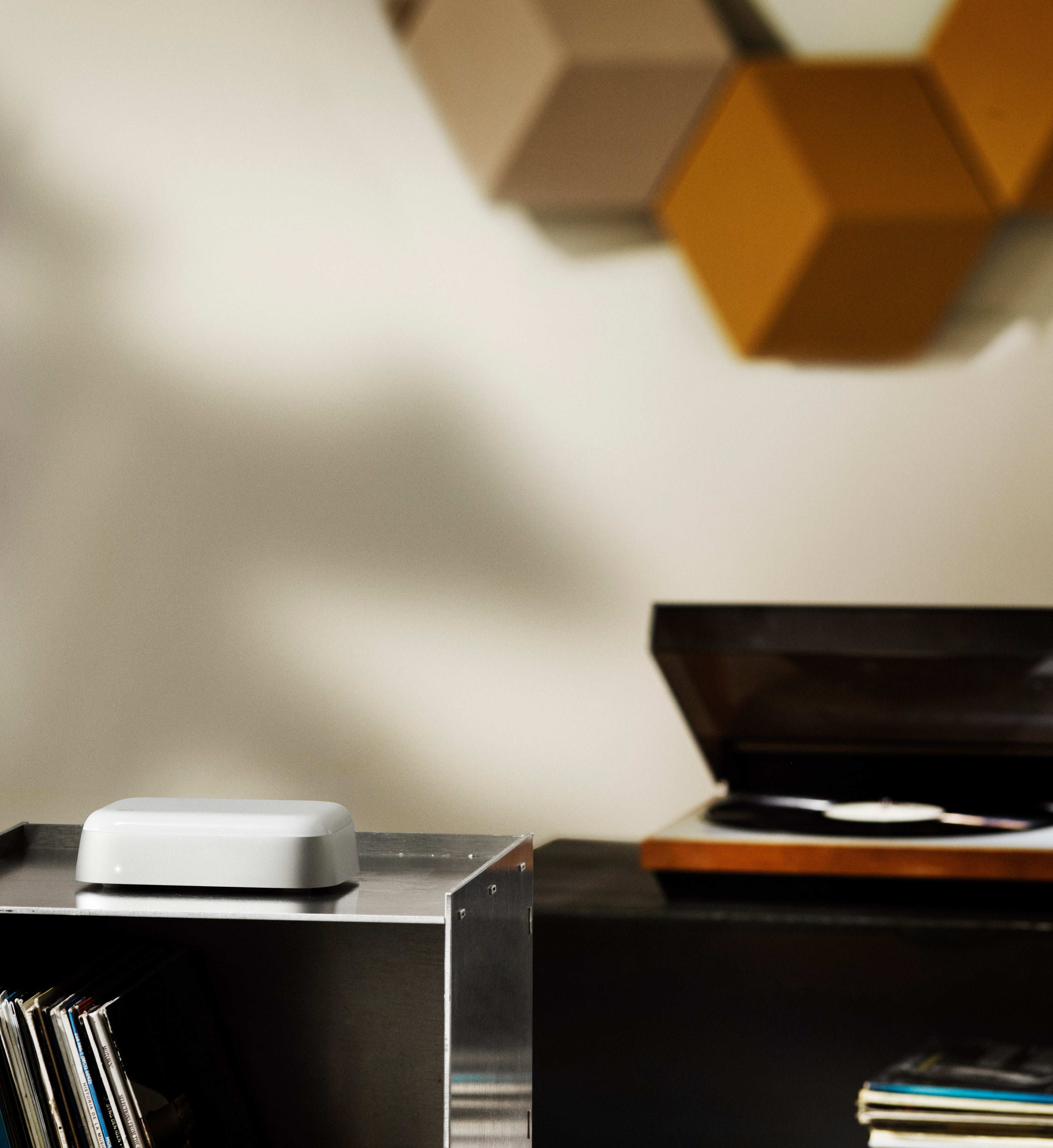 Lifestyle-Ansicht des Bang & Olufsen Beoconnect Core in einem anspruchsvollen Audio-Setup. Das Gerät steht auf einem silbernen Metallregal neben einer Vinyl-Sammlung und einem Plattenspieler. Im Hintergrund sind die modularen Beosound Shape Wandlautsprecher erkennbar, was die Verbindung von analogem Klang und moderner Streaming-Technologie zeigt.