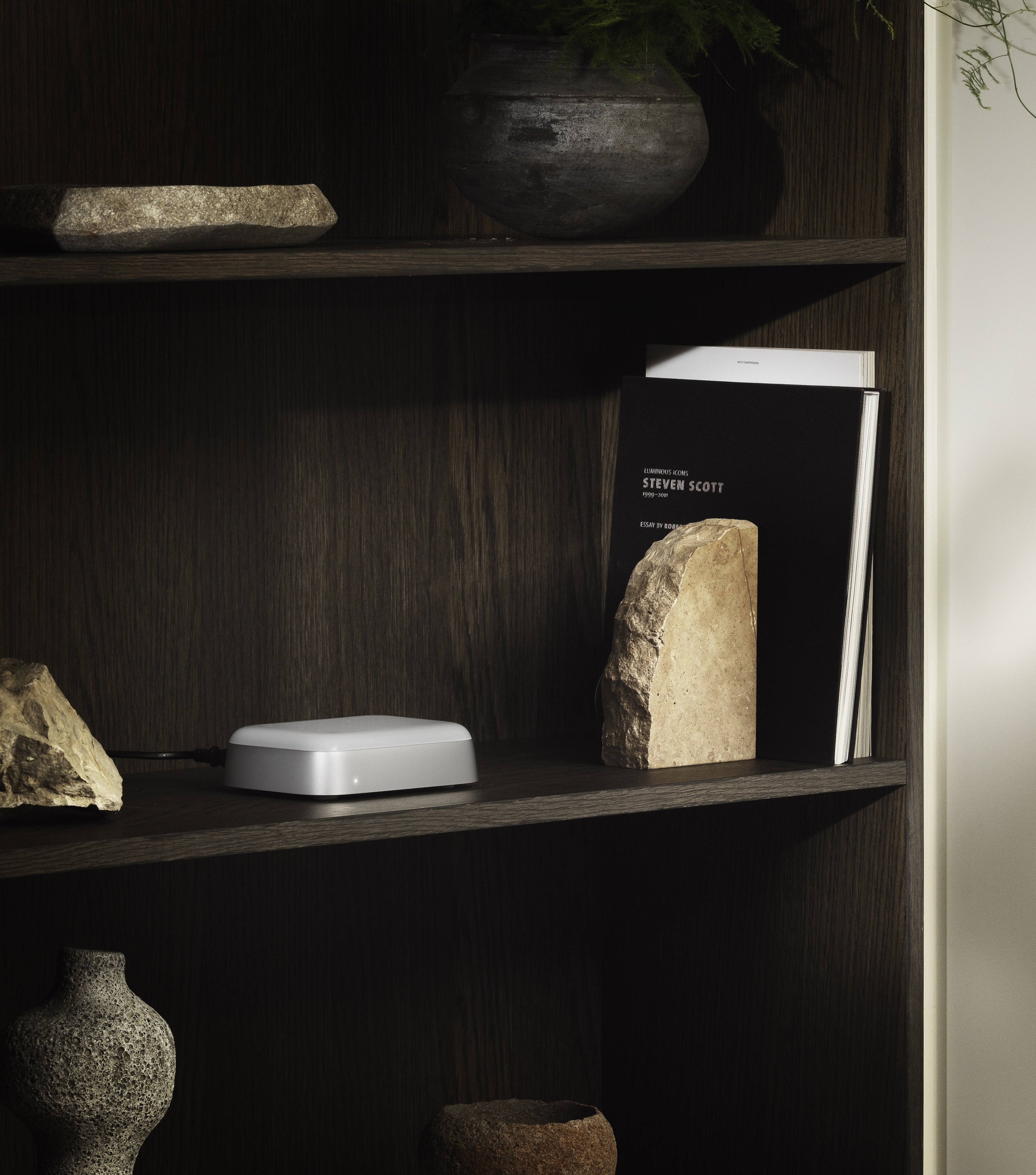 Dezente Lifestyle-Aufnahme des Bang & Olufsen Beoconnect Core Streaming-Hubs. Das kompakte Gerät in Silber und Weiß ist unauffällig in einem dunklen Holzregal zwischen Büchern und dekorativen Steinen platziert, um die nahtlose Integration von smarter Streaming-Technologie in das Interieur zu demonstrieren.