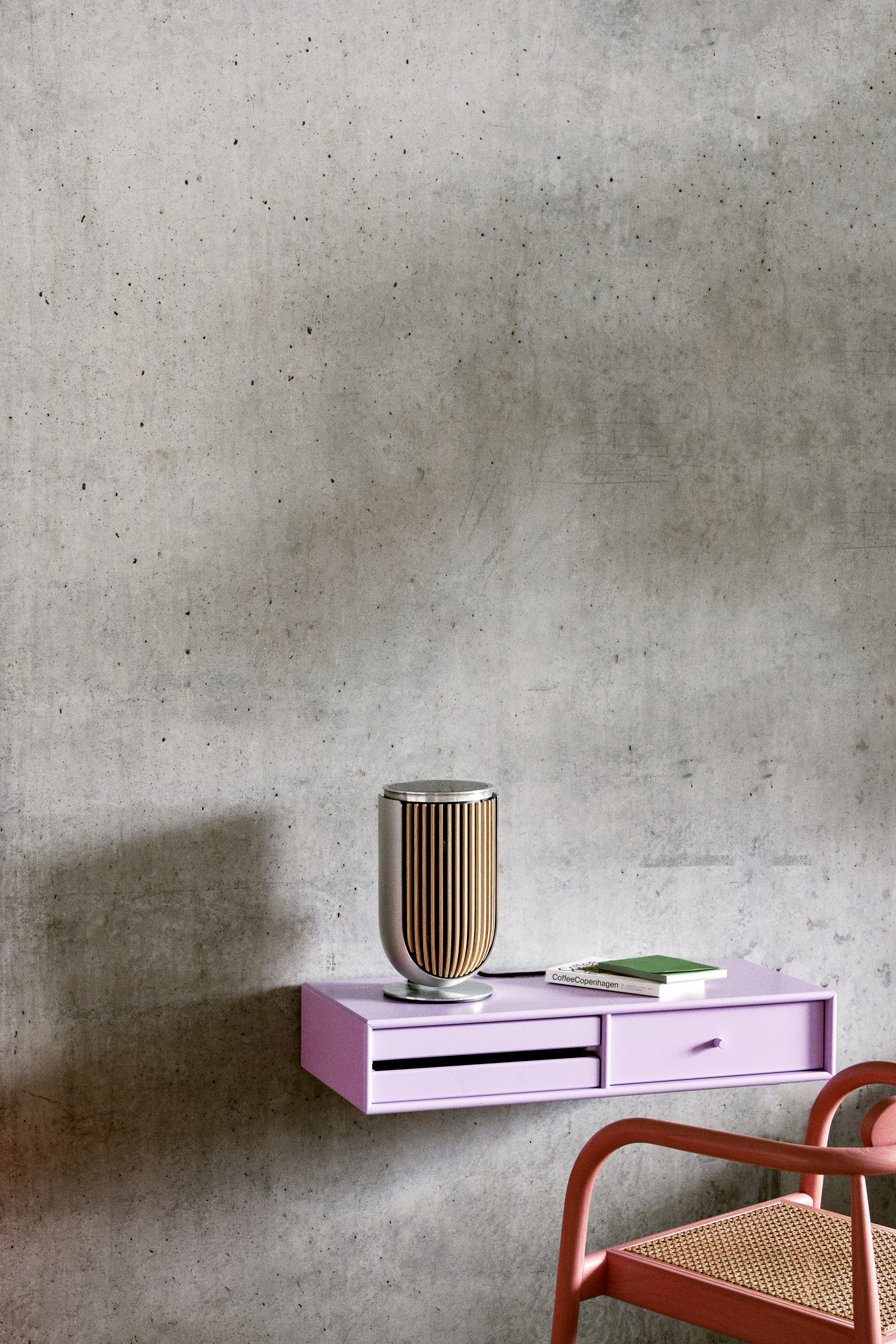 Minimalistische Lifestyle-Aufnahme des Beolab 8 Lautsprechers in Natural Aluminium mit Eichenholz-Lamellen. Der Lautsprecher steht auf einem fliederfarbenen Wandregal vor einer Sichtbetonwand und demonstriert seine kompakte Eleganz als Solitär-System.
