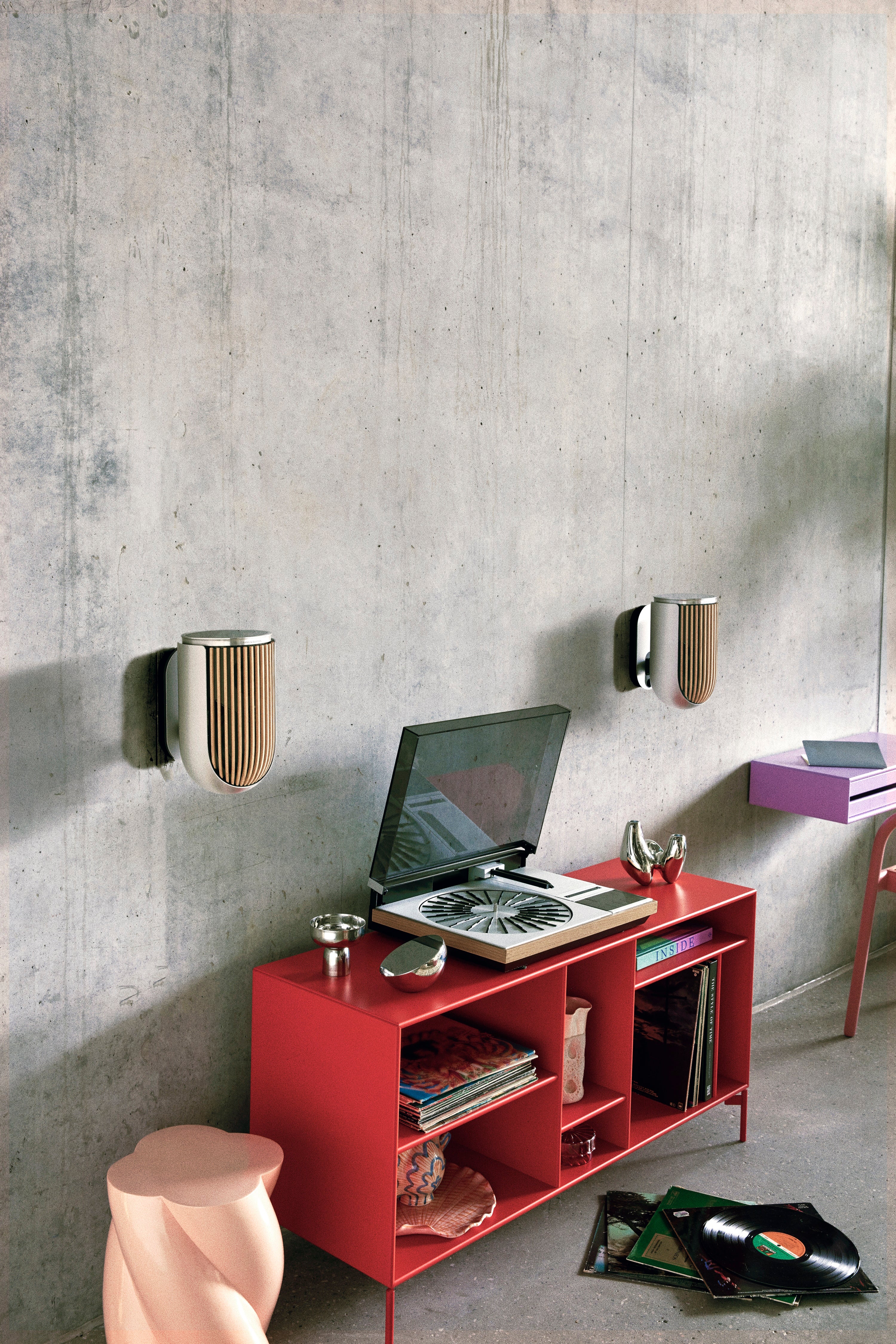 Zwei Bang & Olufsen Beolab 8 Lautsprecher in Natural Aluminium und Eiche, montiert mit diskreten Wandhalterungen an einer Sichtbetonwand. Das Setup wird durch ein rotes Sideboard mit einem klassischen Beogram Plattenspieler ergänzt, was die Symbiose aus analogem Vinyl-Genuss und moderner, platzsparender Audiotechnik zeigt.