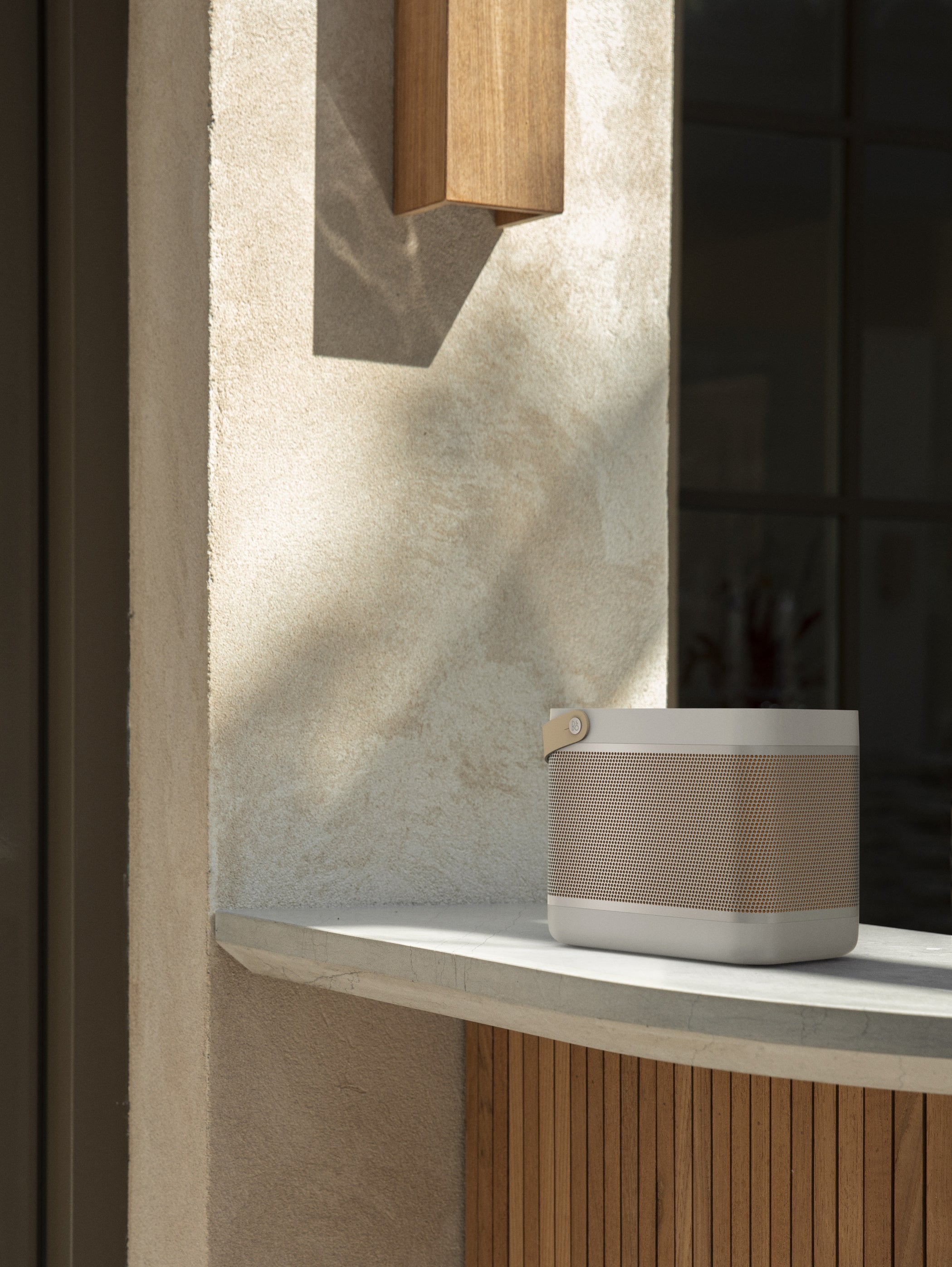 Lifestyle-Aufnahme des Bang & Olufsen Beolit 20 in Grey Mist. Der leistungsstarke portable Bluetooth-Lautsprecher steht auf einem Sims vor einer hellen Putzwand. Das Bild betont das eloxierte Aluminium-Gitter, den markanten Trageriemen aus pflanzlich gegerbtem Leder und die integrierte kabellose Qi-Ladefläche auf der Oberseite.