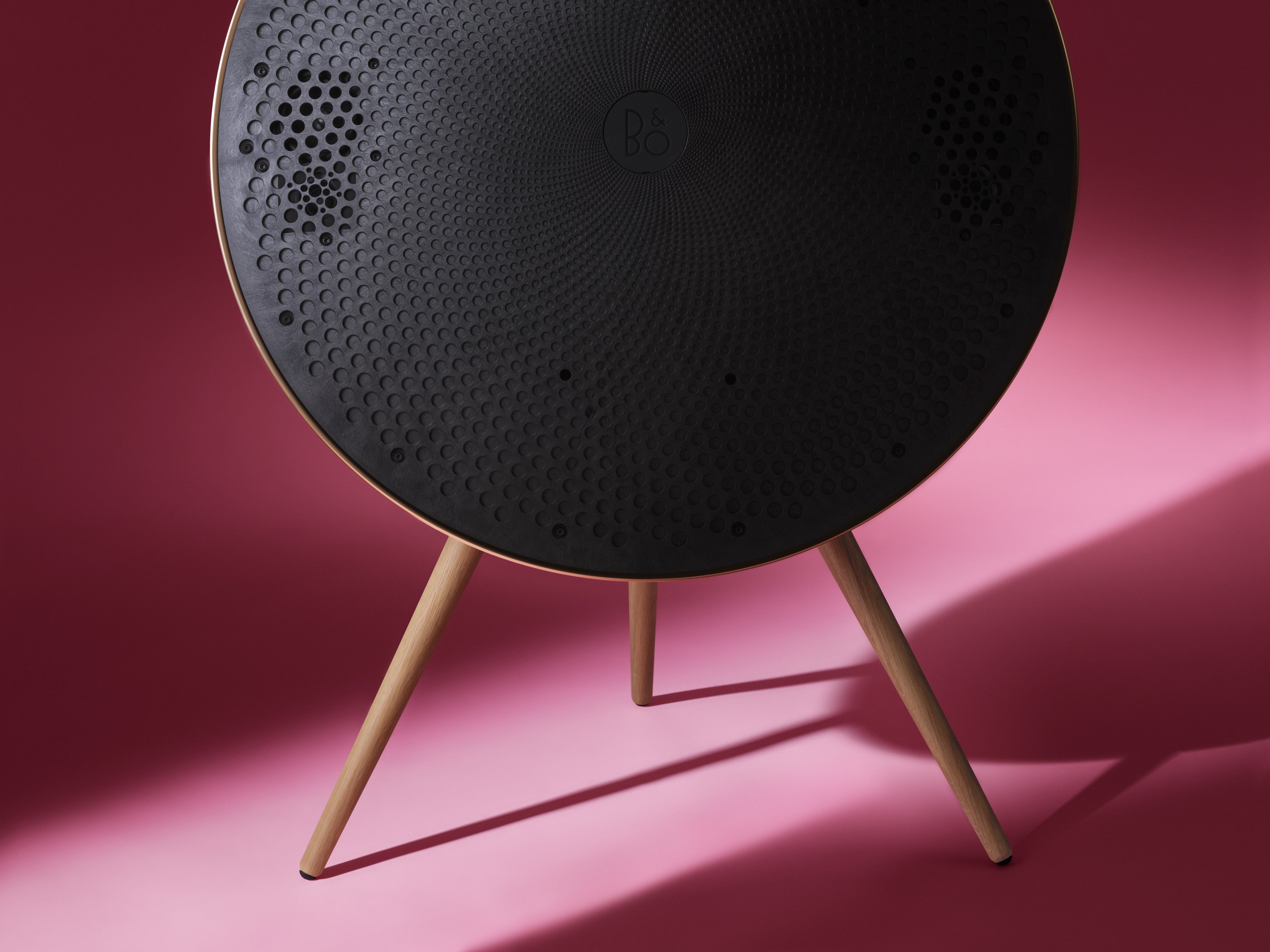 Nahaufnahme der Bang & Olufsen Beosound A9 ohne Stoffabdeckung. Die Ansicht zeigt die interne Treiber-Anordnung und die akustische Perforation der Frontschale. Der goldfarbene Aluminiumring und die Eichenholzbeine bilden einen luxuriösen Kontrast zum markanten, pinkfarbenen Hintergrund.