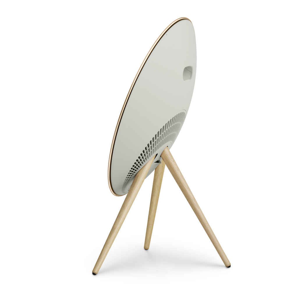 Dreiviertel-Ansicht der Rückseite einer Bang & Olufsen Beosound A9 in Gold Tone. Das Bild betont das fließende, weiße Gehäuse-Design, den integrierten Tragegriff und die ästhetisch gestalteten Belüftungsöffnungen. Die Beine aus heller Eiche vervollständigen den hochwertigen Möbel-Look.