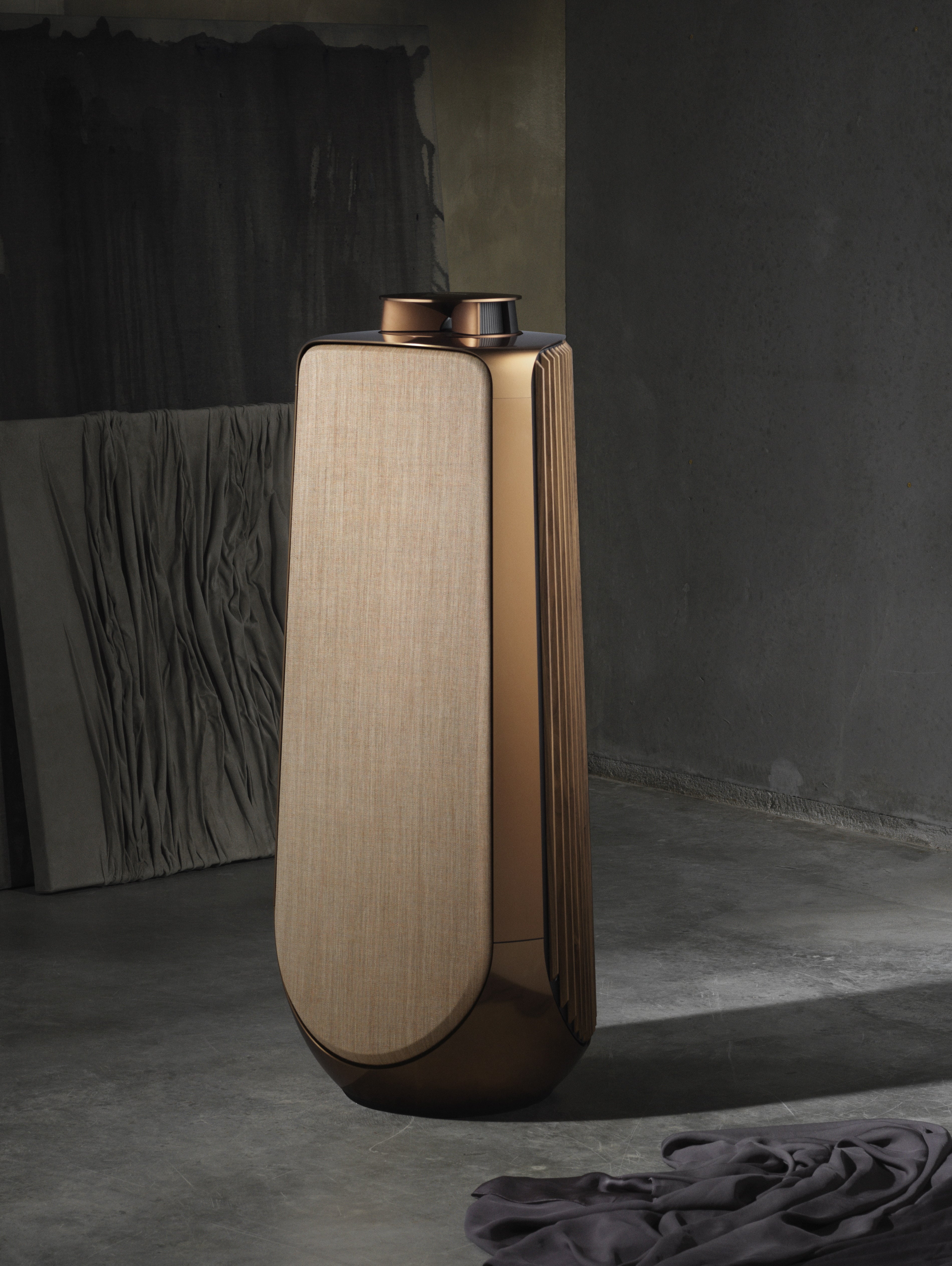 Skulpturale Studioaufnahme des Bang & Olufsen Beolab 50 High-End-Aktivlautsprechers in Bronze Tone. Die Ansicht zeigt die Kombination aus poliertem, bronzefarbenem Aluminium, warmen Eichenholz-Lamellen und hochwertigem Textilbezug vor einem minimalistischen, industriellen Hintergrund mit dramatischem Lichtspiel.