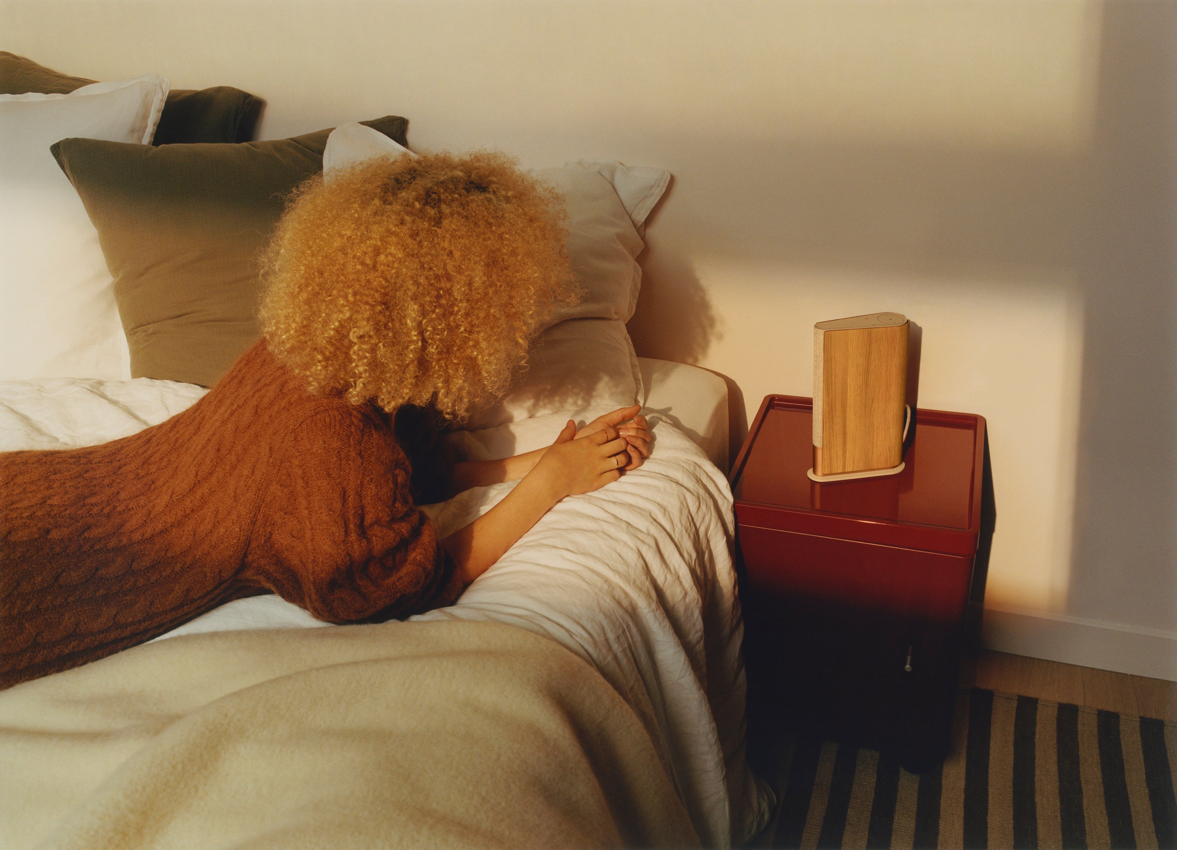 Warme Lifestyle-Aufnahme eines Bang & Olufsen Beosound Emerge Lautsprechers in Gold Tone, der als Nachttisch-Lautsprecher auf einer tiefroten Kommode steht. Eine Frau liegt entspannt auf dem Bett im Vordergrund, während sanftes, spätnachmittägliches Sonnenlicht die Szene erhellt.
